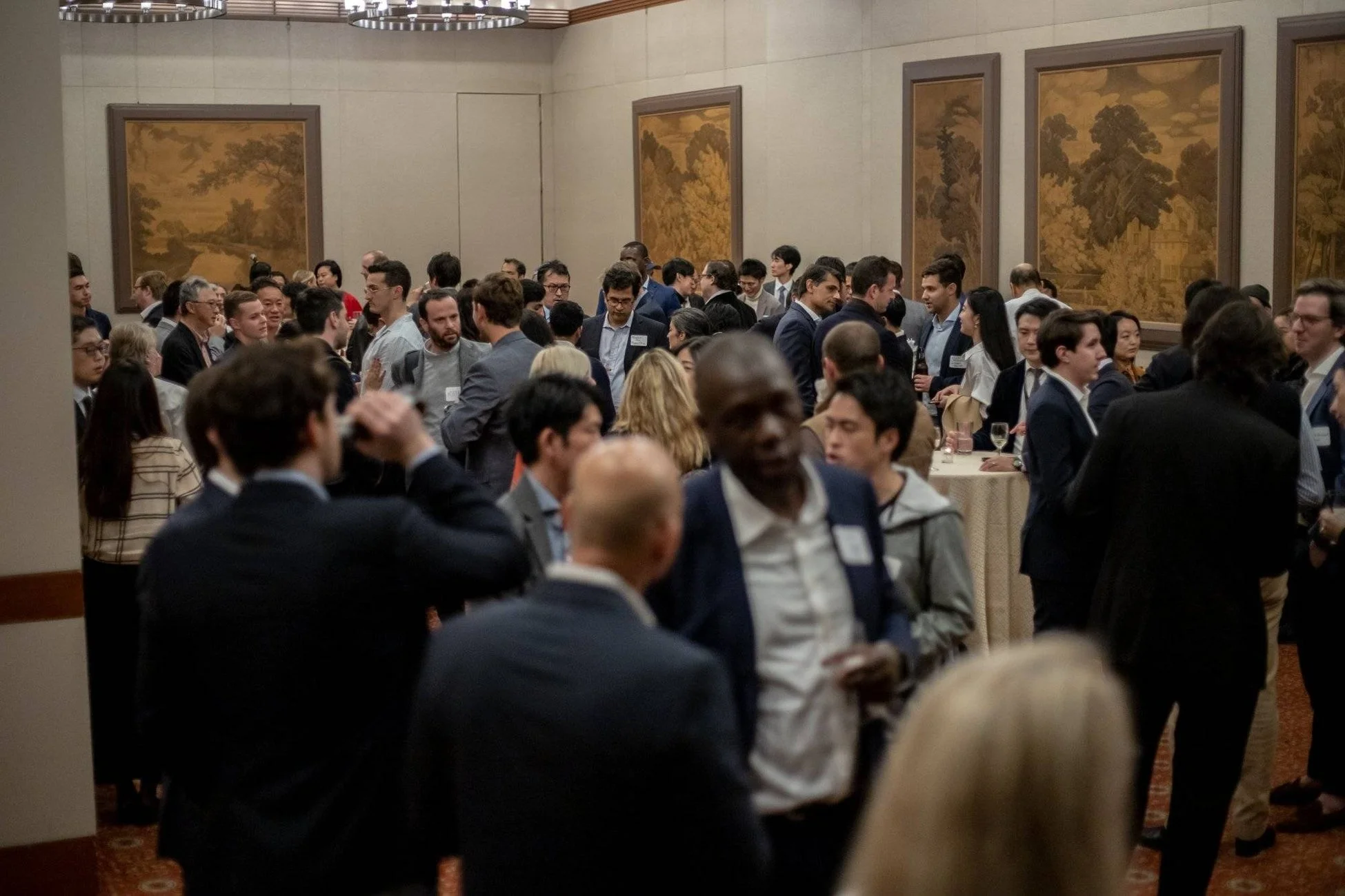 多くの人々が集まって交流している屋内のイベントの様子です。参加者はビジネスカジュアルな服装で、談笑したり飲み物を片手に過ごしており、壁には絵画が飾られています。