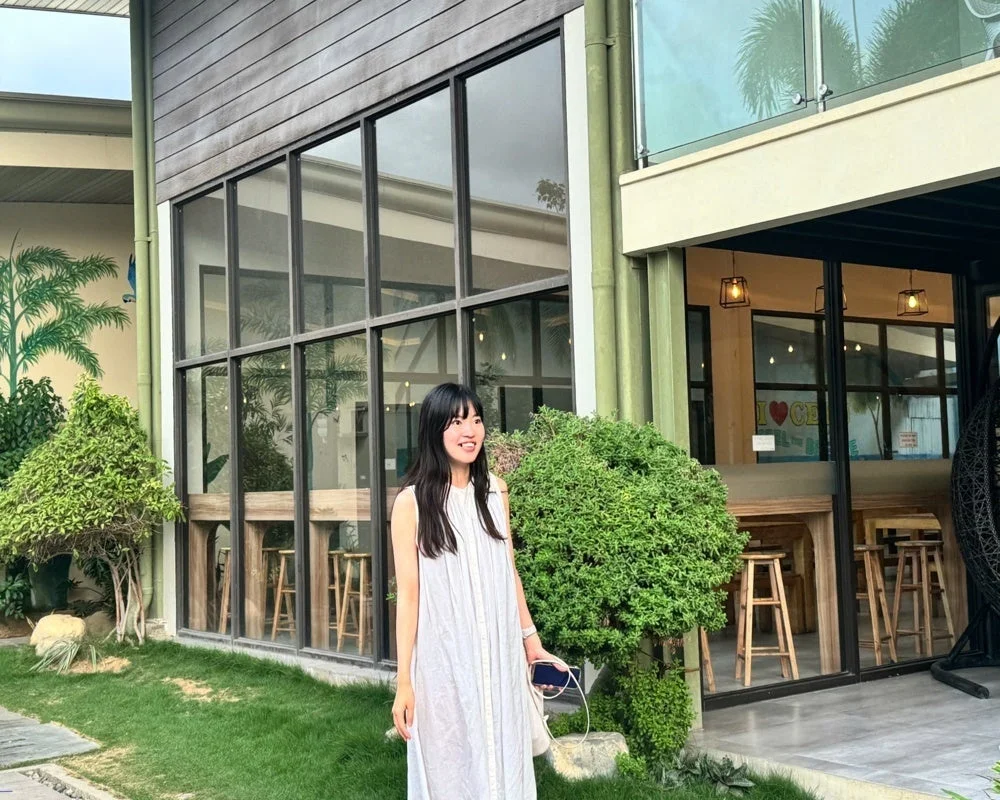 白いワンピースを着た女性が、大きな窓のあるモダンな建物の前にある緑豊かな庭で笑顔で立っています。カフェのような空間が窓越しに見え、全体的に穏やかな雰囲気です。