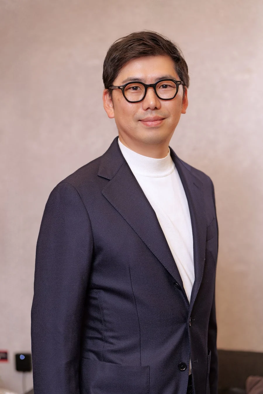 ダークスーツに白いタートルネック、眼鏡をかけた東洋系の男性がカメラに向かって微笑んでいるポートレート写真です。