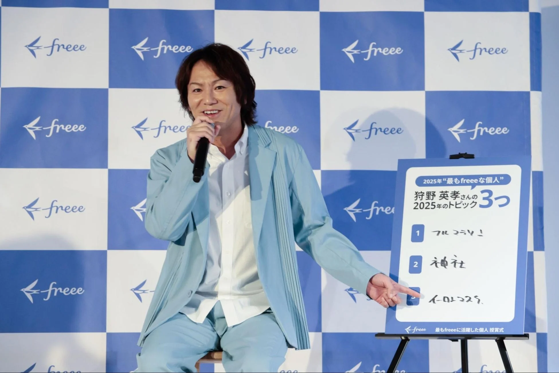 狩野英孝さんがfreeeのイベントで、ライトブルーのスーツ姿でマイクを持ち、2025年の目標について書かれたボードを指しながら笑顔で話している様子。freeeのロゴが背景に広がる授賞式の風景。