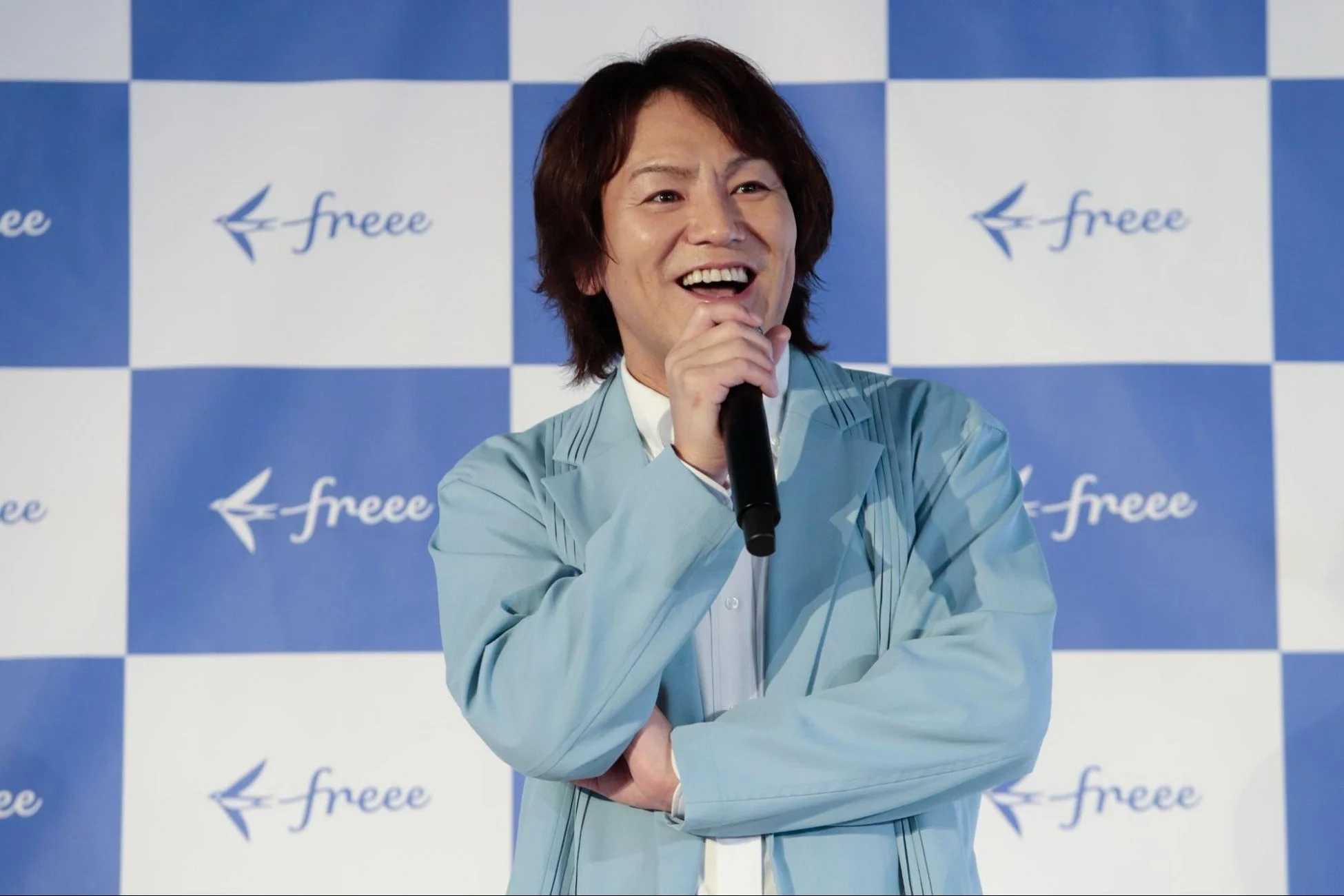 水色のジャケットを着た男性がマイクを持ち、笑顔で話している様子。背景には「freee」のロゴが繰り返しデザインされたチェック柄のパネルがある。
