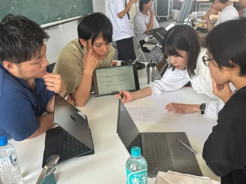 数人の男女がテーブルを囲み、ノートパソコンやタブレットを使って共同作業や議論をしているリーダー合宿の様子。