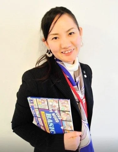 笑顔でカメラを見つめるアジア人女性が、スーツとスカーフを着用し、英語の絵辞書を抱えています。