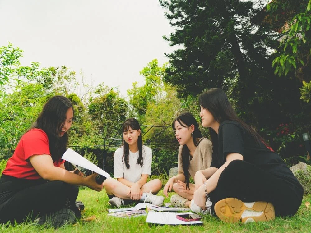 4人の若い女性が屋外の芝生の上に座り、書類を囲んで会話をしたり、勉強したりしている