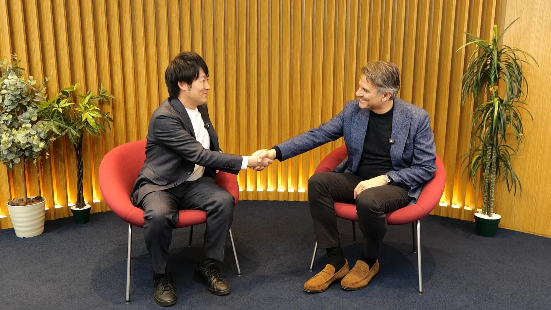 2人の男性が赤い椅子に座り、笑顔で握手をしている様子