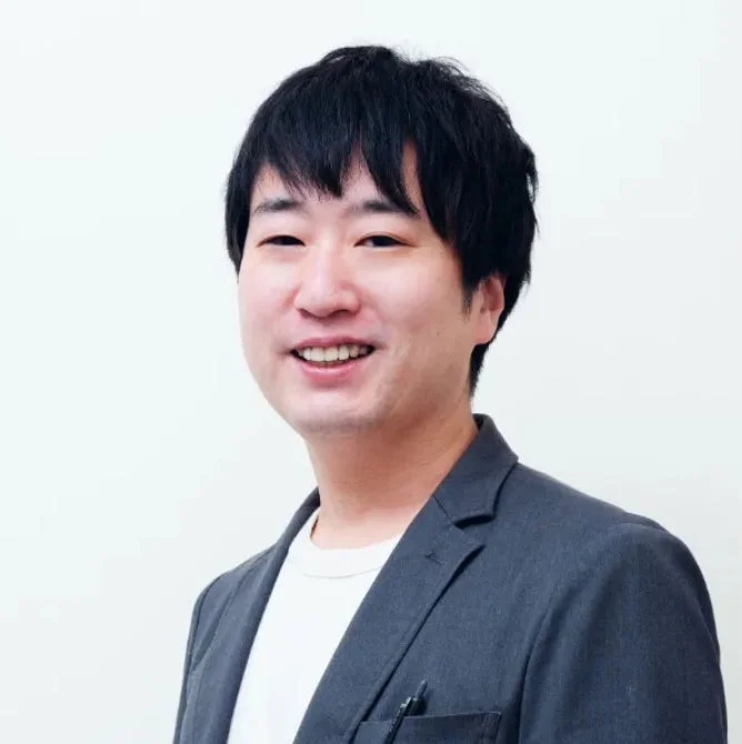 白い背景の前で笑顔を見せるアジア人男性のポートレート。黒髪で、濃いグレーのジャケットと白いTシャツを着用しており、親しみやすい印象を与えます。