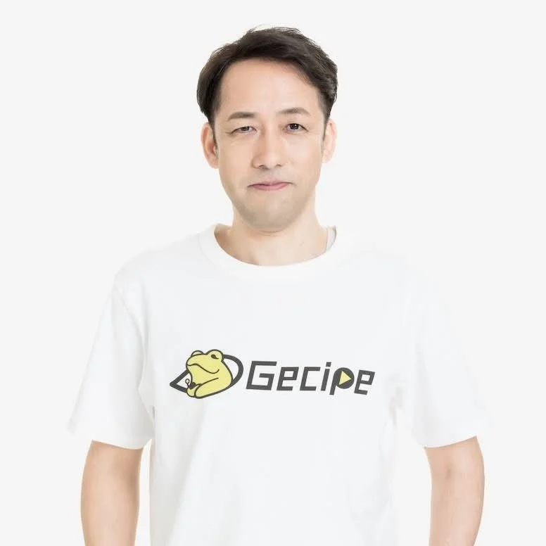 白いTシャツを着た男性がカメラに向かって立っています。Tシャツにはカエルのキャラクターと「Gecipe」という文字のロゴが描かれています。