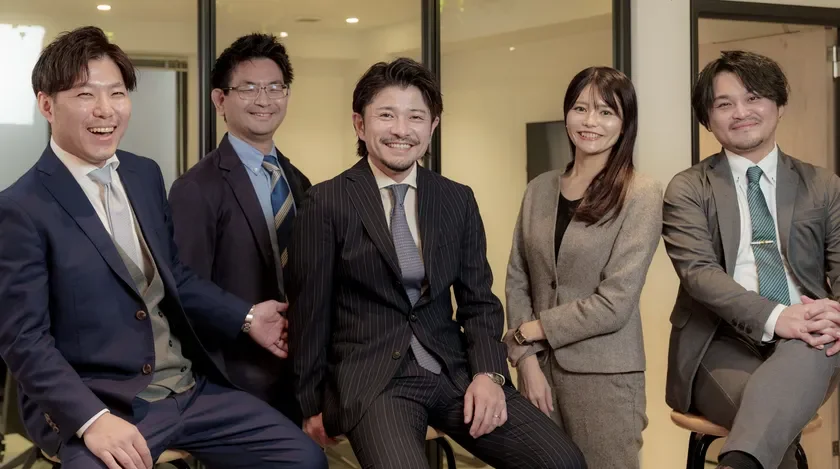 オフィスでスーツを着用した5人のビジネスパーソンが笑顔で座っている集合写真。