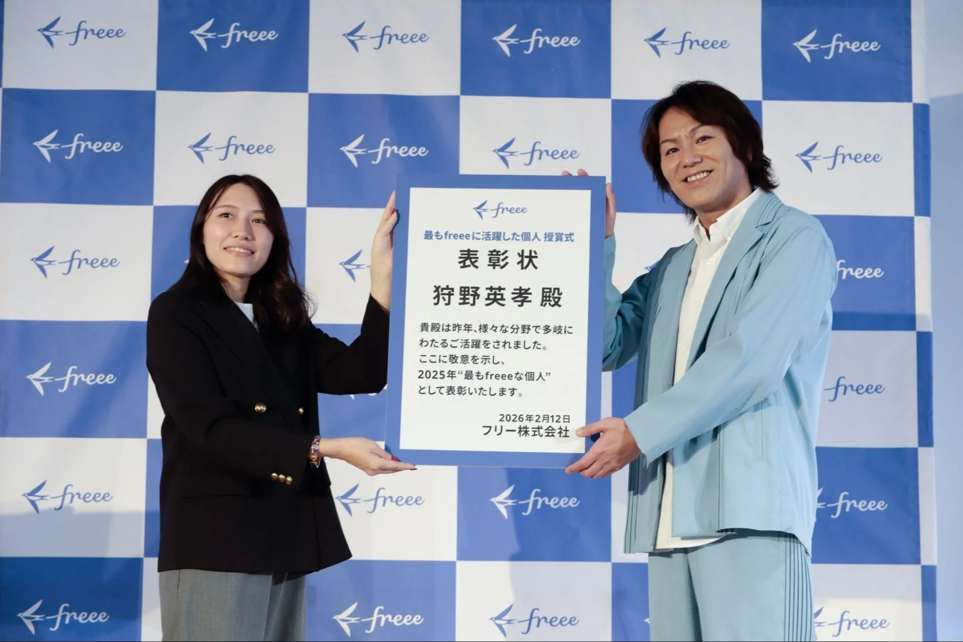 freee株式会社のロゴが背景に広がる会場で、女性がタレントの狩野英孝さんに表彰状を授与している様子です。狩野英孝さんは2025年の「最もfreeeな個人」として表彰されています。