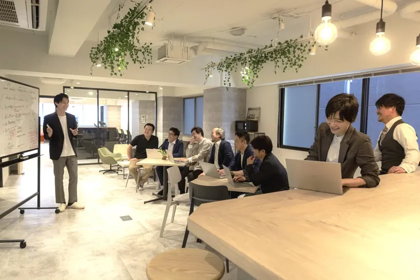 会議室で一人の男性がホワイトボードを使ってプレゼンテーションを行っており、他のメンバーは笑顔で話を聞いている。