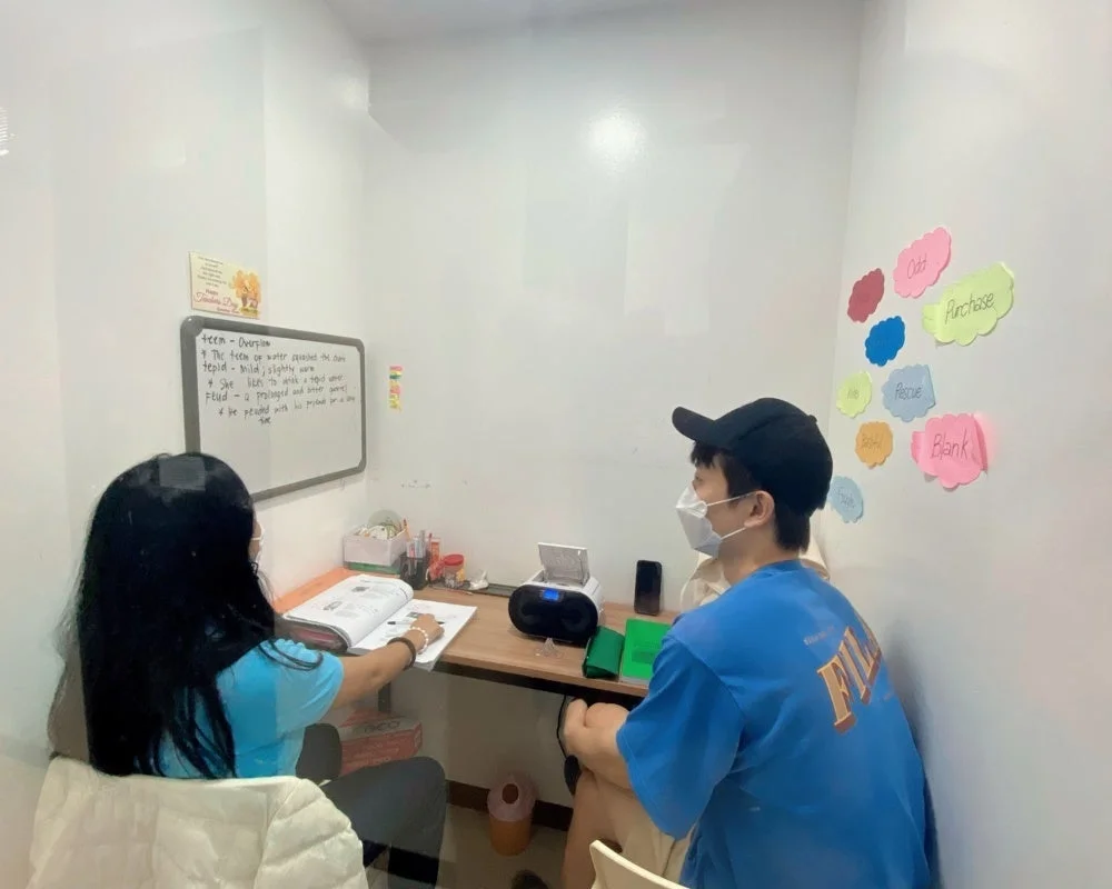 白い部屋で、マスクを着用した生徒と先生らしき人物が机に向かい、英語を学習しています。ホワイトボードには英単語の定義があり、壁には単語が書かれた付箋が貼られています。