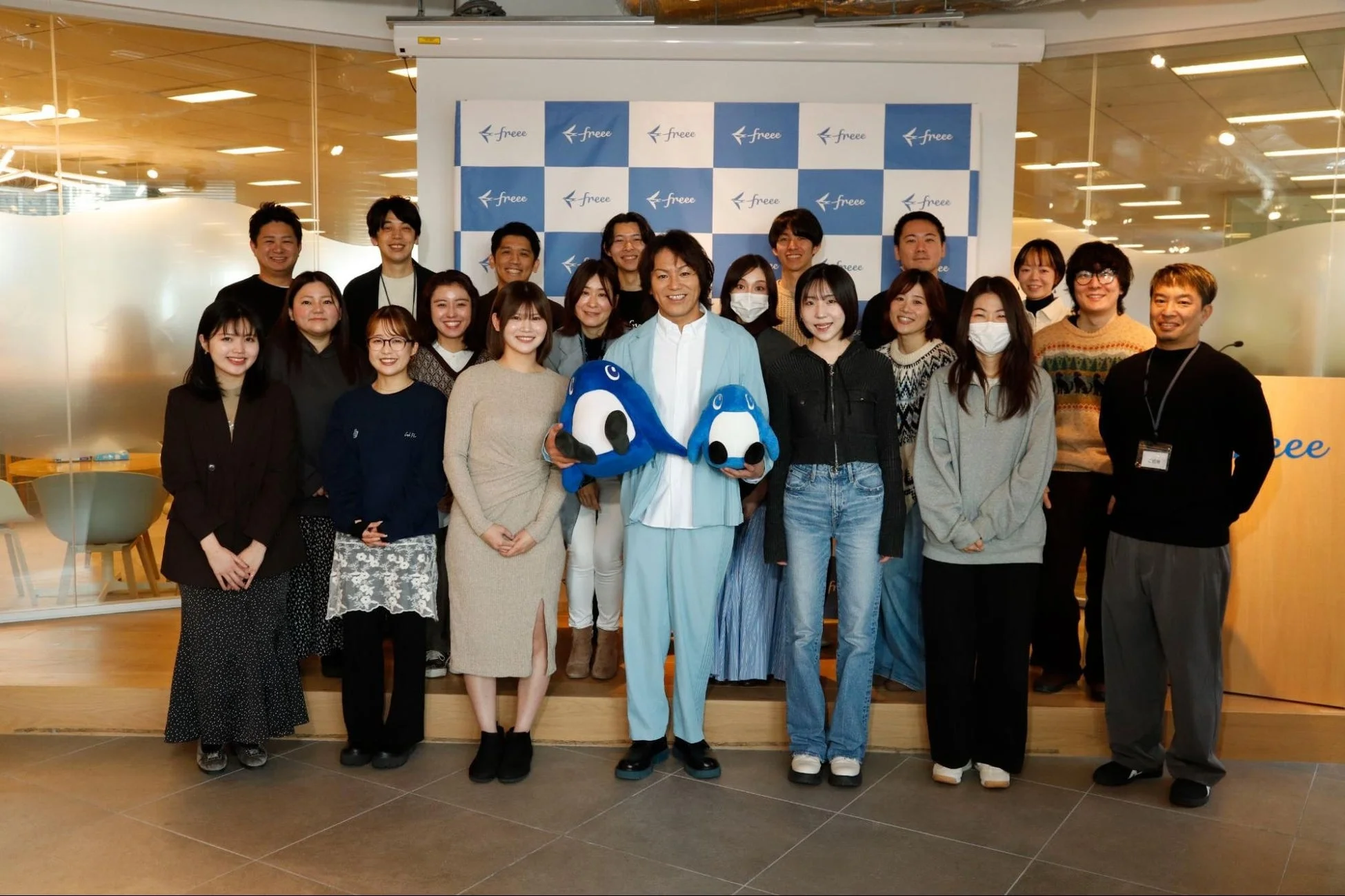 オフィス内で撮影された、freeeのロゴが入った背景の前で笑顔を見せる大勢の従業員の集合写真です。中央の男性は青いペンギンのようなマスコットを2つ持っています。