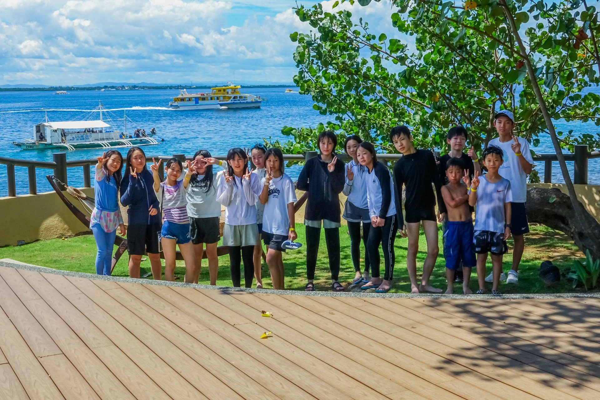 青い海と空を背景に、木製デッキでポーズをとる子供たちとティーンエイジャーのグループ写真です。数隻のボートが浮かび、南国でのアイランドツアーを楽しんでいる様子がうかがえます。