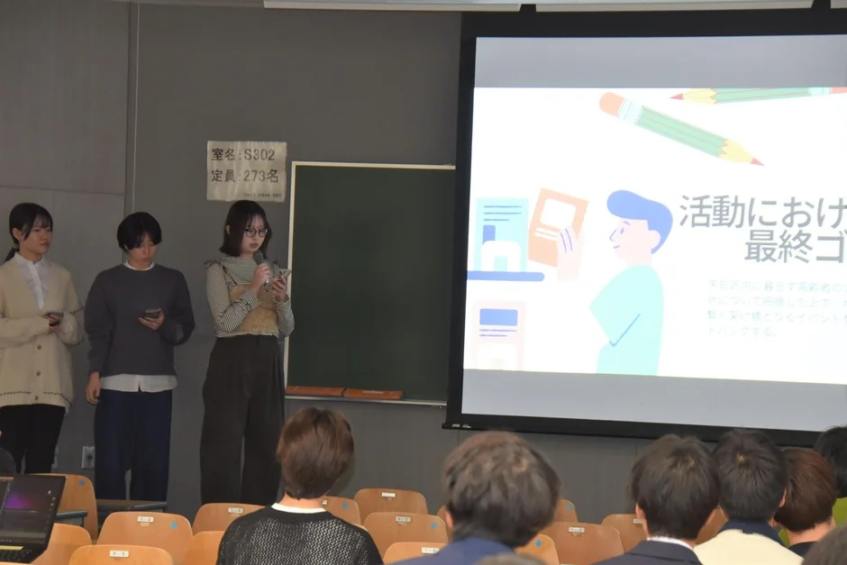 大学の教室で、3人の学生がプロジェクターを使ってプレゼンテーションを行っています。中央の学生がマイクを持ち発表しており、多くの聴衆が耳を傾けている様子です。