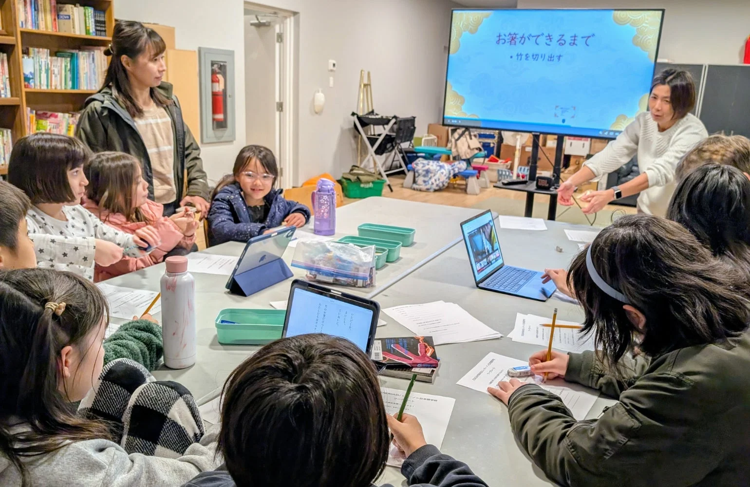 日本語学校の教室で、子供たちがタブレットやノートパソコンを使いながら熱心に学習している様子です。スクリーンには「お箸ができるまで」というテーマが表示され、女性講師が指導しています。グループで学ぶ活気ある教育風景が捉えられています。