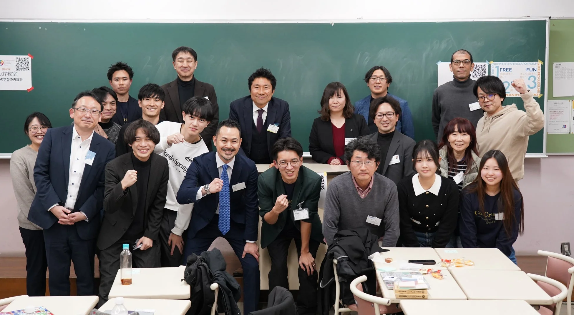教室の黒板を背景に、学生と大人を含む多数の日本人男女が笑顔で集合写真に収まっている