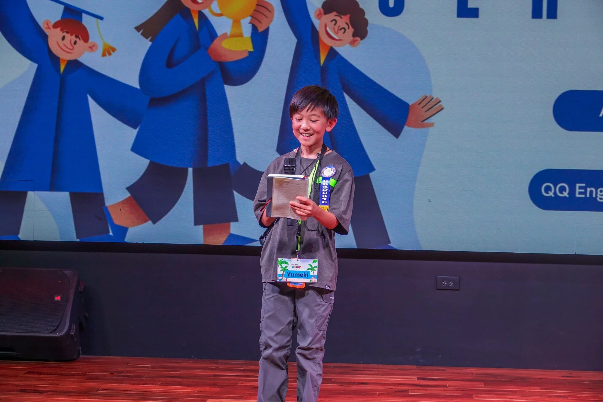 笑顔の少年がステージ上でノートを手に持ち、発表している様子。背景には卒業ガウンを着たキャラクターのアニメーションが映し出されており、卒業イベントか表彰式のような雰囲気である。