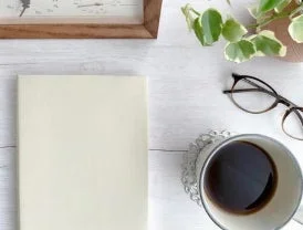 白い木目のデスクに、無地のノート、温かいコーヒー、メガネ、観葉植物が配置されたフラットレイの画像です