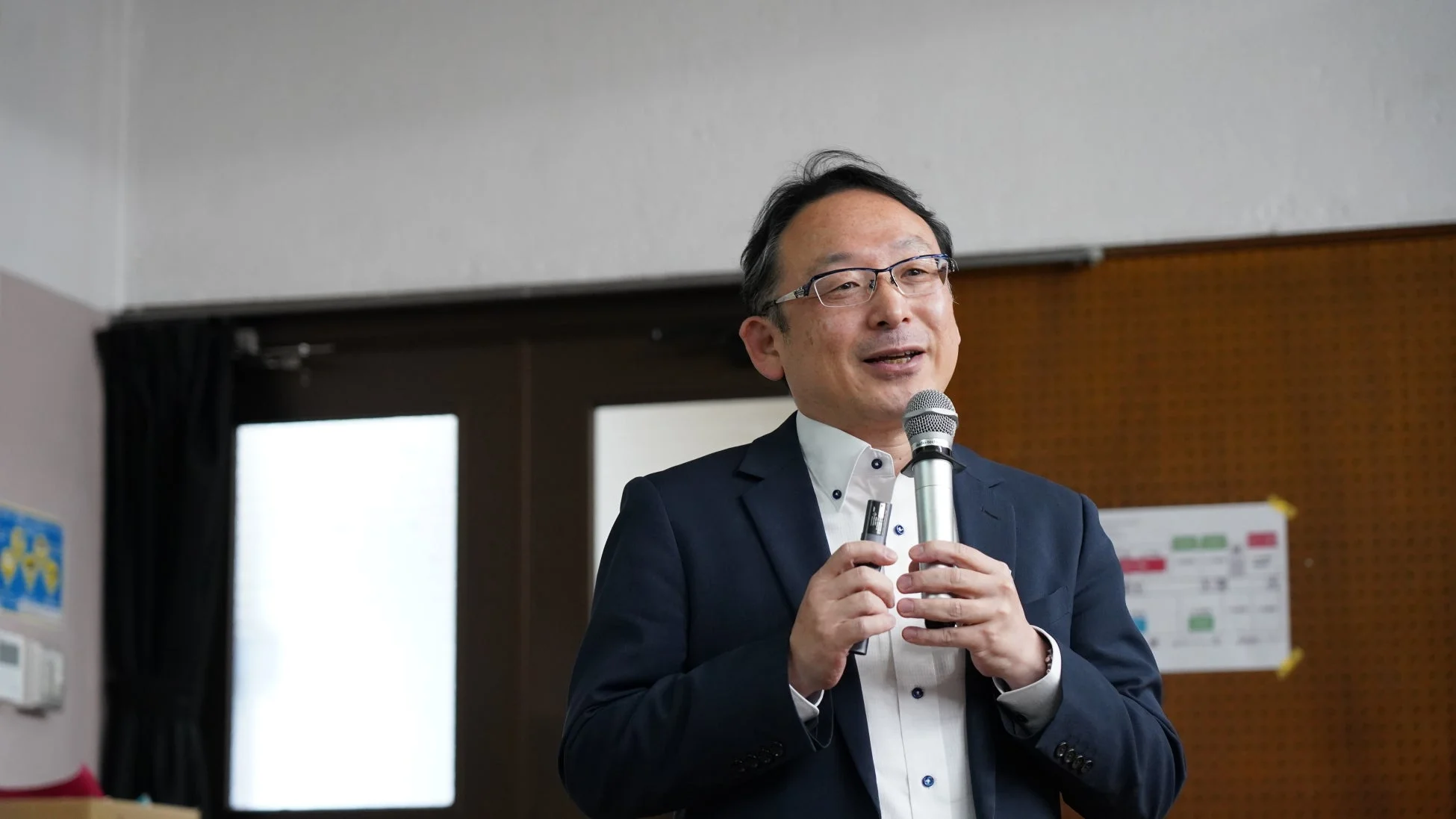 紺色のスーツと眼鏡をかけた男性がマイクを片手に笑顔で話しています