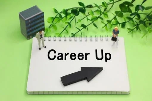 ノートに「Career Up」という文字と上向きの矢印が描かれ、その周りにビジネスマンのミニチュア人形が配置されています