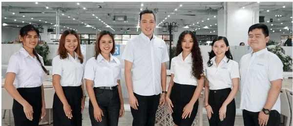 明るいオフィス環境で、白シャツと黒いボトムスの制服を着た男女7人が笑顔でポーズをとっています。チームや職場の集合写真のようです。