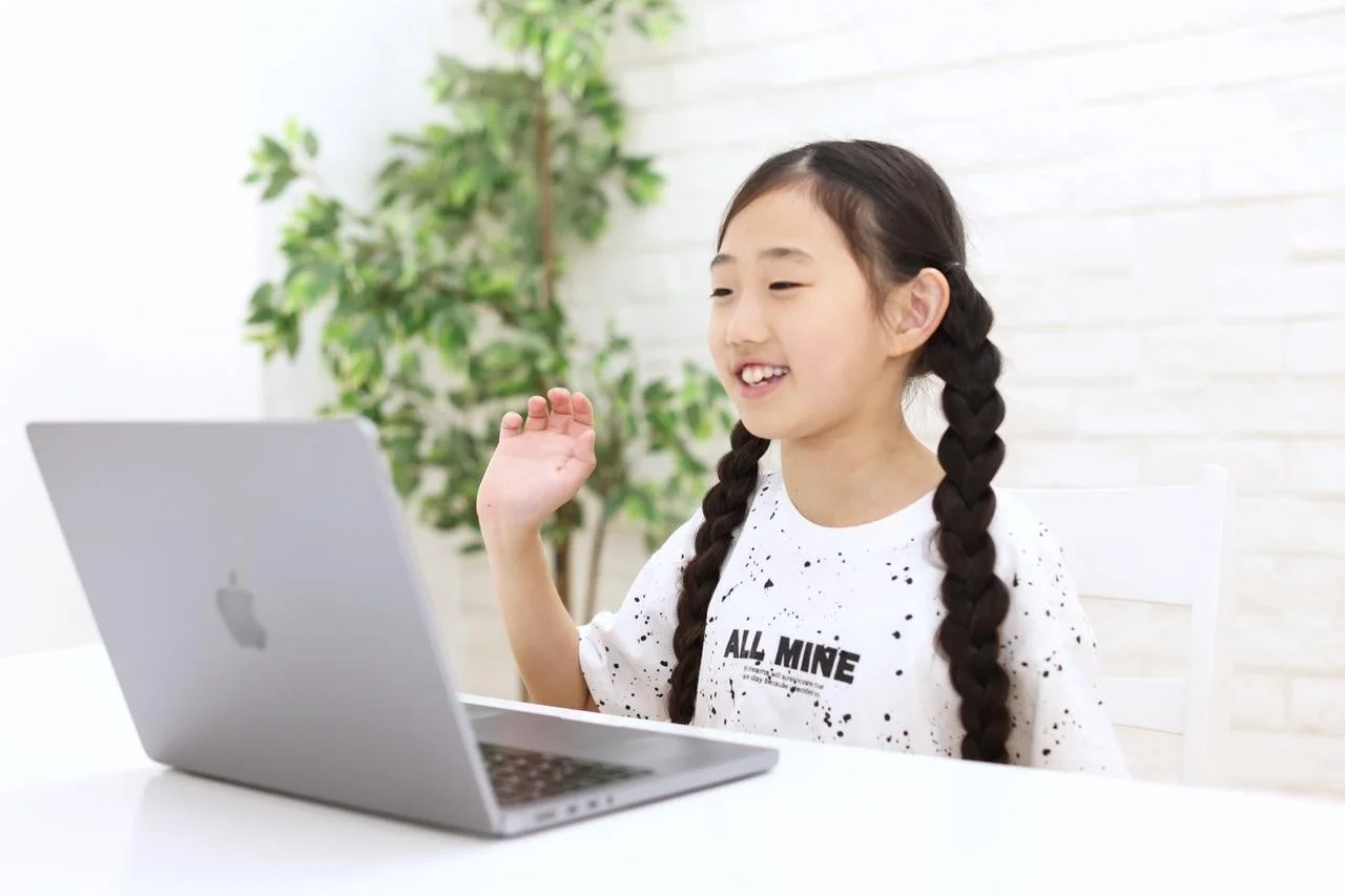 笑顔の女の子がノートパソコンに向かい、手を振っています。オンラインでのコミュニケーションや学習の様子を捉えた一枚です。