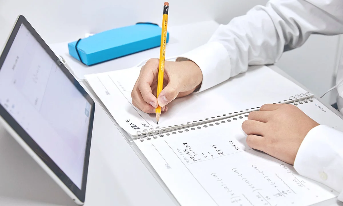 白いシャツを着た人物が、ノートに鉛筆で数学の問題を解いています。左手前にはタブレットが置かれており、集中して学習に取り組む様子がうかがえます。