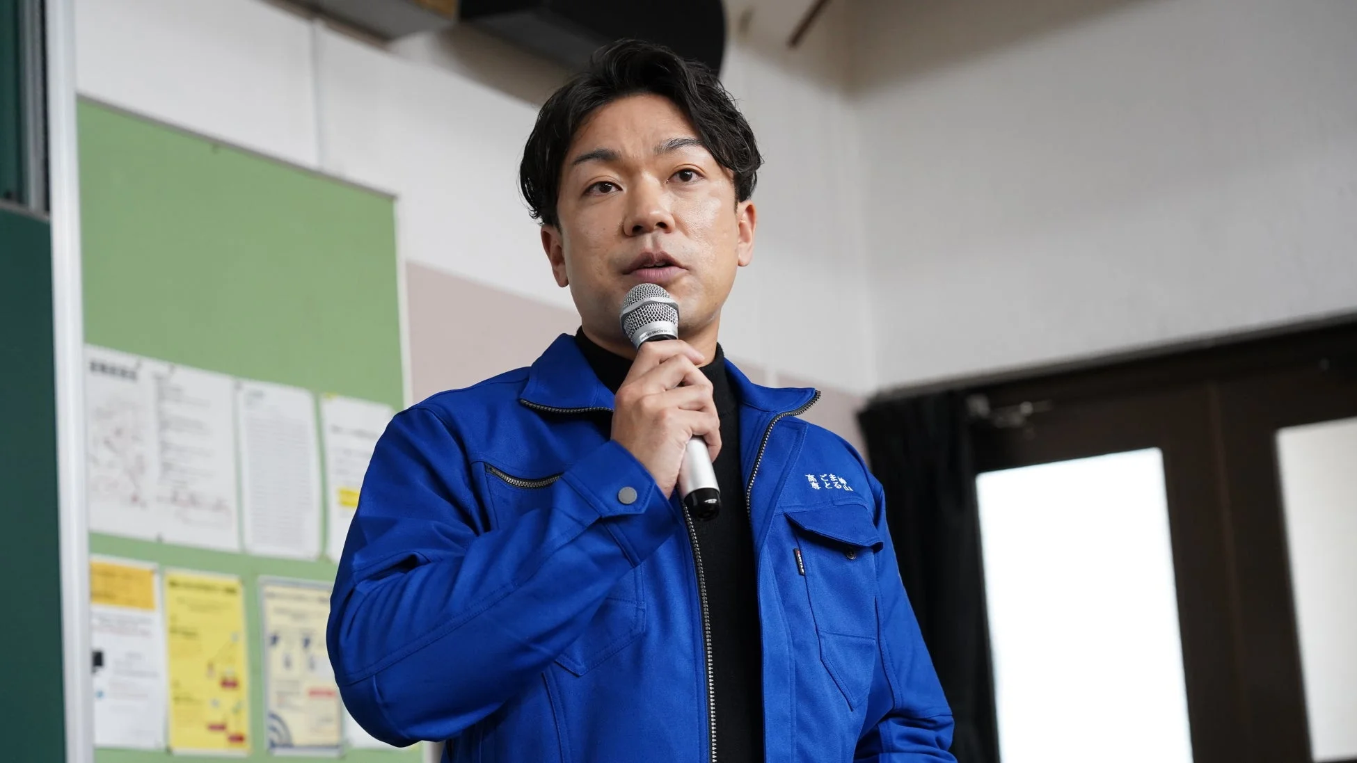 青い作業着を着た男性が、屋内でマイクを手に持ち、真剣な表情で話している様子です