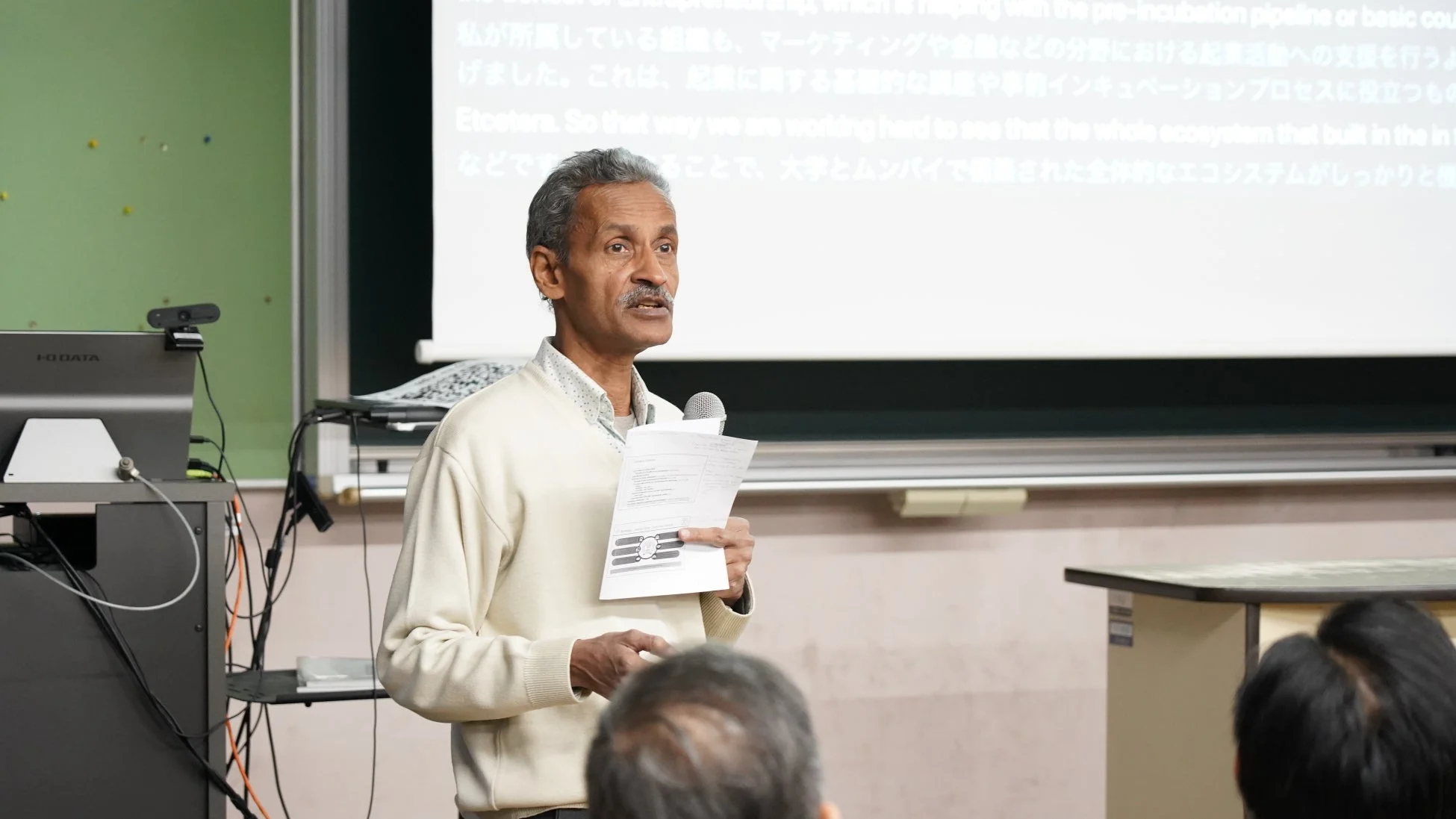 講演会場で、一人の男性が聴衆に向けてプレゼンテーションを行っています