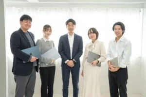 5人のビジネスパーソンが室内でノートパソコンを持ち、笑顔で並んで立っている集合写真