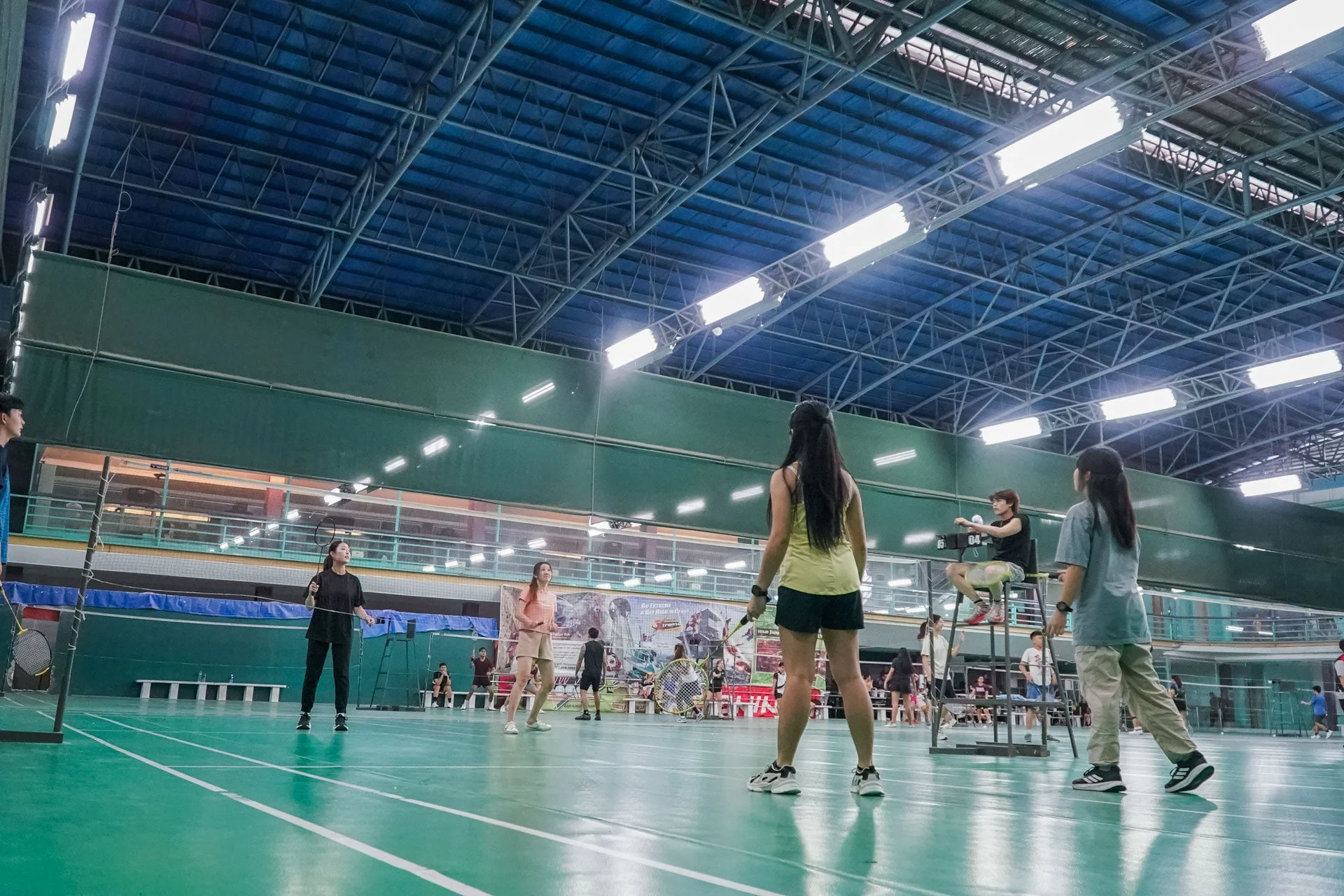 体育館のバドミントンコートで、数人がバドミントンを楽しんでいる様子。審判台には審判が座っており、活気あるスポーツの瞬間が捉えられています。