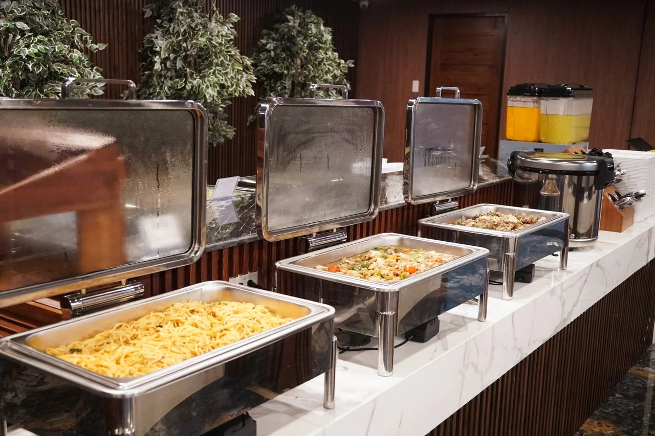 ホテルやレストランのビュッフェ形式の食事風景です。数種類の温かい料理が並べられたチェーフィングディッシュには、パスタや炒め物のような料理が見えます。飲み物や食器も用意されており、食欲をそそる場面です。