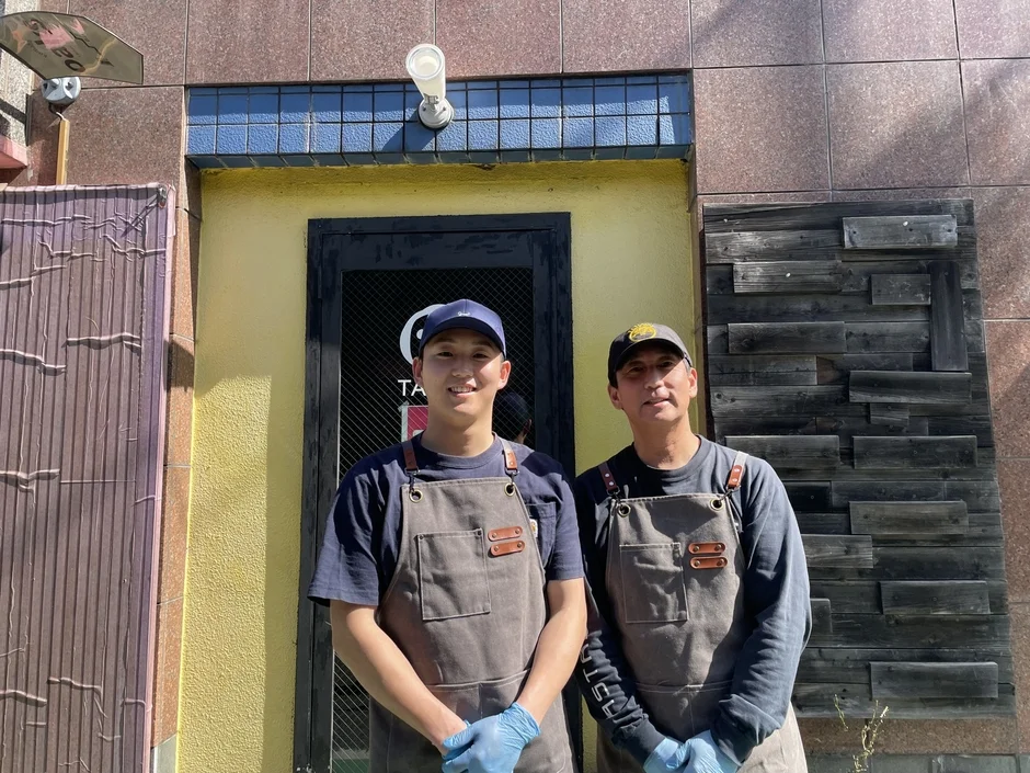 エプロンと帽子を着用した2人の男性が、黄色い店舗の前に立っている