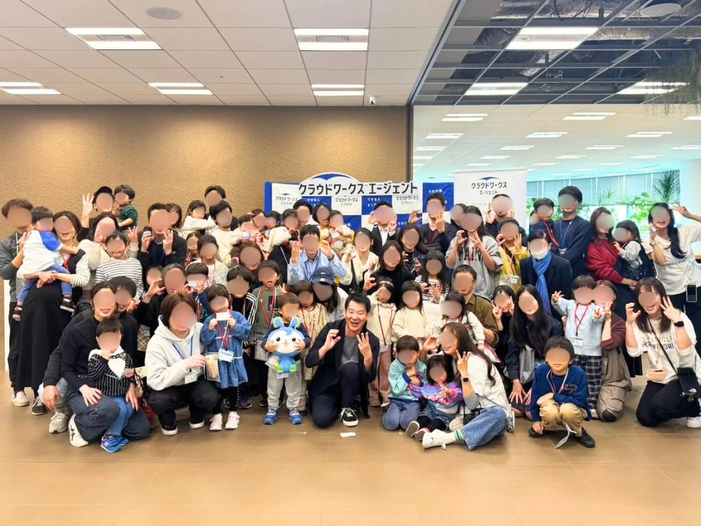 屋内で撮影された大人と子供が大勢写る集合写真です。背景には「クラウドワークス エージェント」のバナーがあり、参加者たちは笑顔でポーズをとっています。