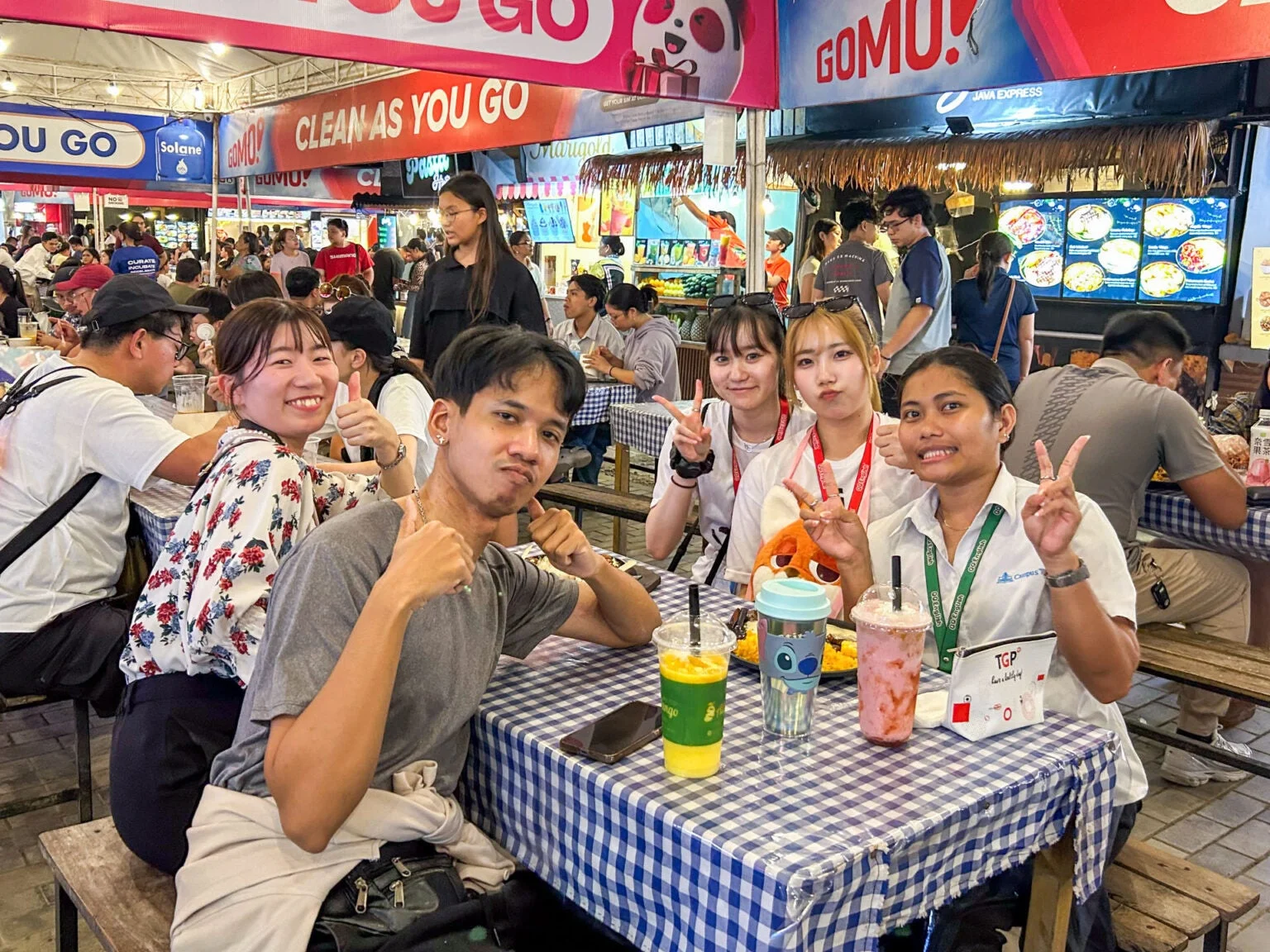 賑やかなフードコートで、友人らしき若者たちが飲み物を片手に楽しそうに過ごしている様子。皆笑顔で、サムズアップやピースサインをしており、和やかな雰囲気が伝わります。