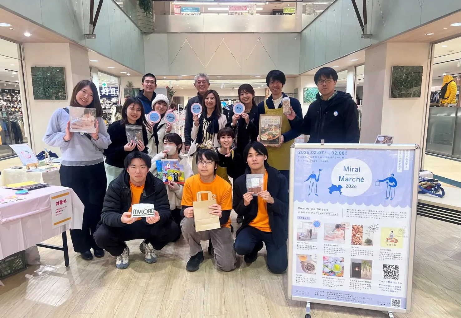 ショッピングモール内で開催される「Mirai Marché 2026」のイベントスタッフや出展者と思われる人々が集合写真を撮っている様子。イベント告知ボードには開催日時や場所、出展者情報が記載されている。