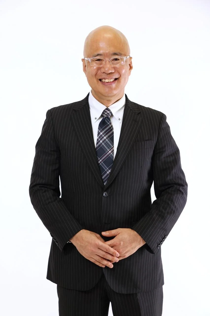 白い背景の前で、黒いストライプのスーツと眼鏡を着用したアジア人男性が、笑顔でカメラを見つめています。両手を前で組んでおり、ビジネスシーンでのポートレート写真です。