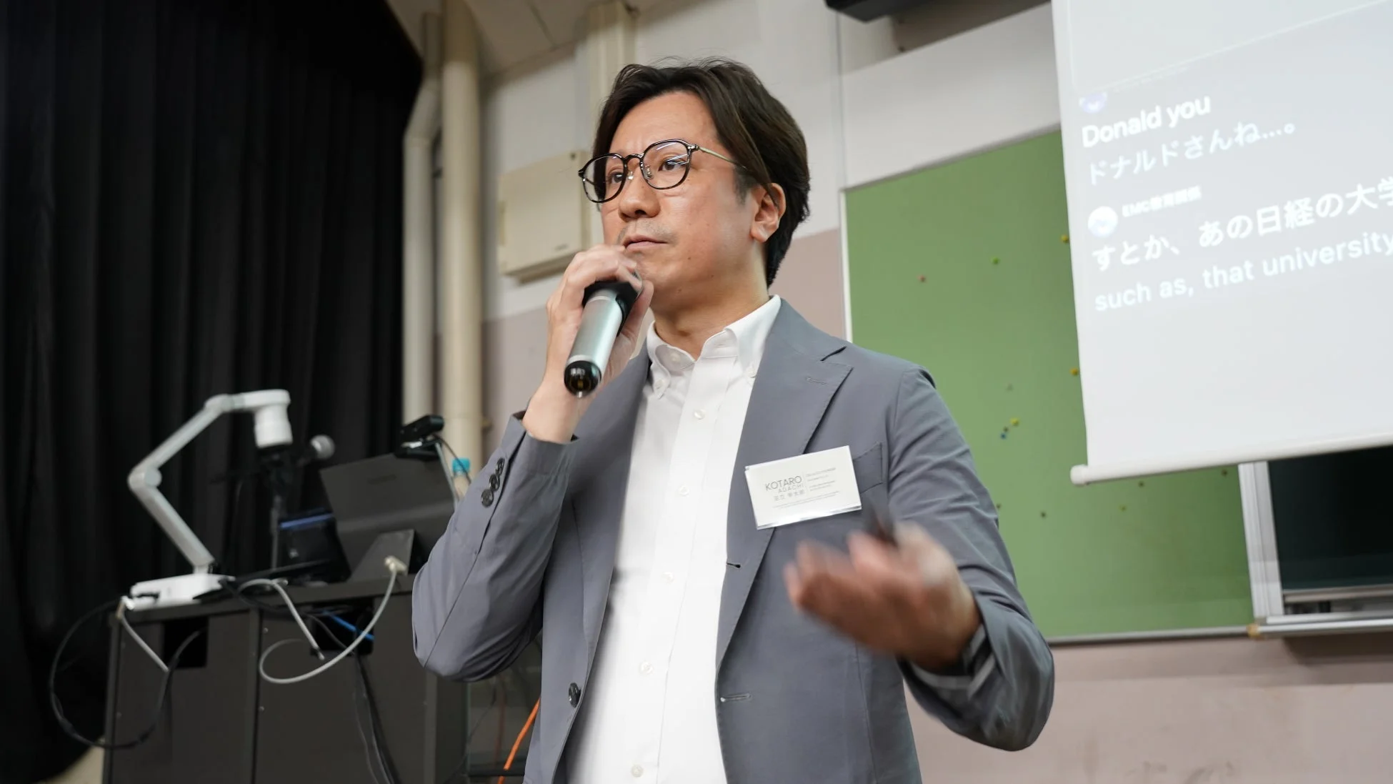 眼鏡をかけた男性がマイクを手に持ち、ステージ上で講演を行っている様子です