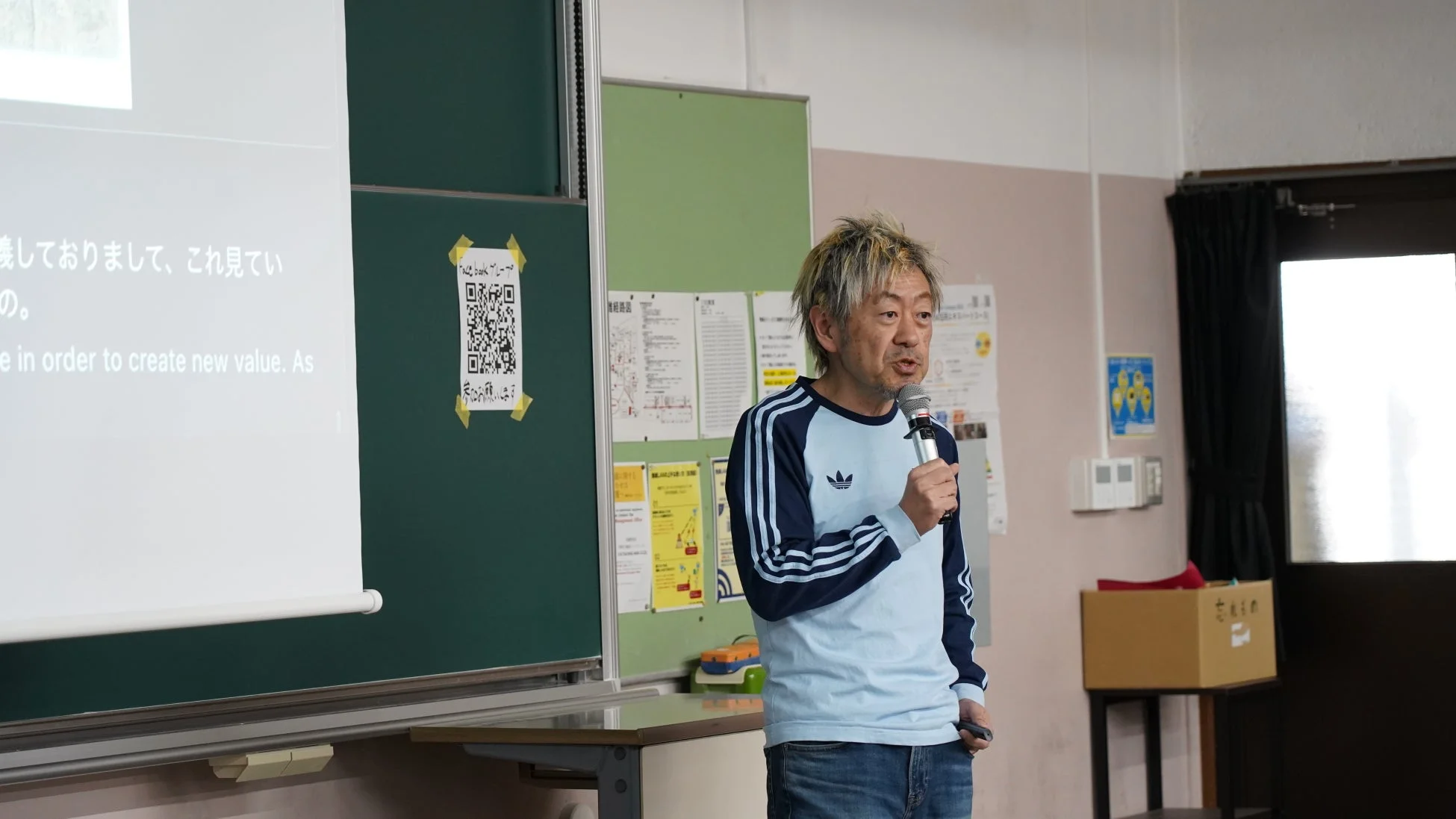 尖った髪型の男性が、教室のような場所でマイクを手にプレゼンテーションを行っています