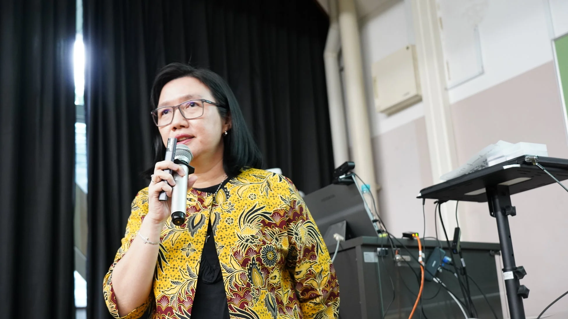 眼鏡をかけた女性が、黄色いバティック柄のジャケットを着用し、マイクを持って話している様子です