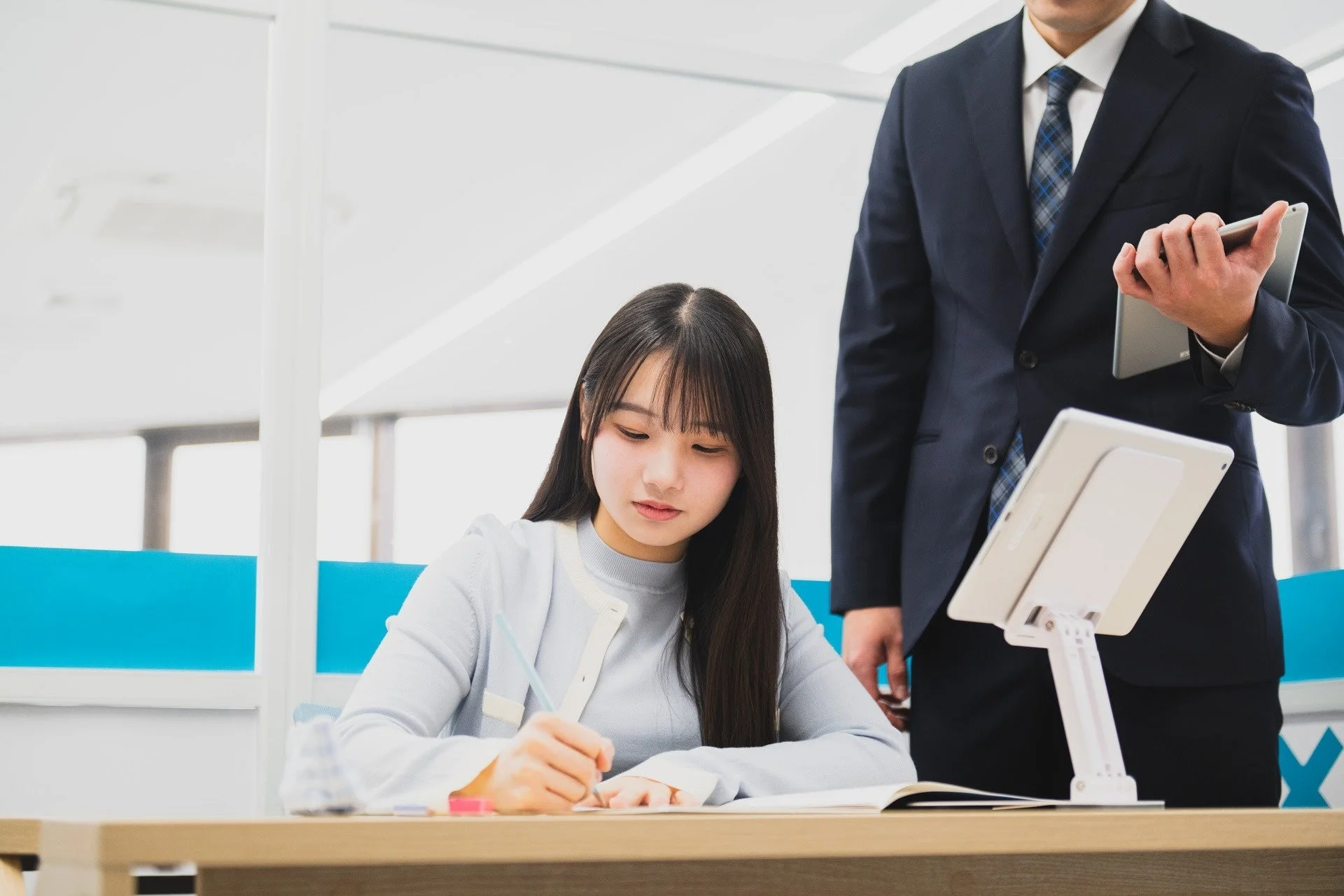 オフィスで、デスクに座ってノートに書き込む若い女性と、その横でタブレットを持つスーツ姿の男性の様子。
