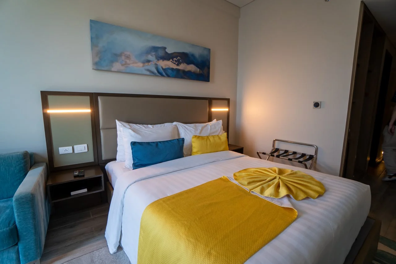 白と黄色のリネンが特徴的なモダンなホテルの一室です。広々としたベッドには青と黄色の枕が置かれ、壁には抽象的なアートが飾られています。清潔で快適な雰囲気が漂っています。