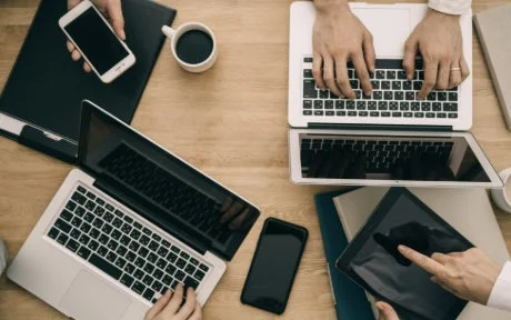 複数の人がノートパソコン、スマートフォン、タブレットといったデジタルデバイスを使い、木製のデスクで共同作業や会議をしている様子を上から捉えた画像です