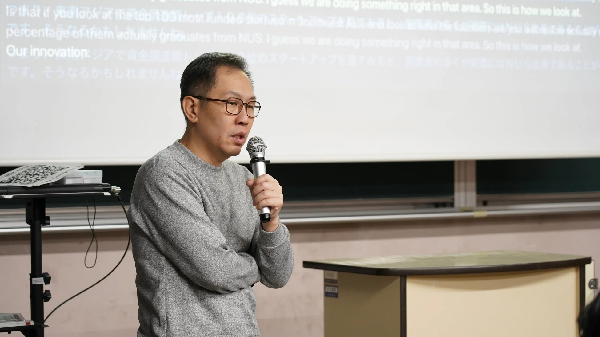 眼鏡をかけたアジア系の男性が、マイクを持って講演している様子です