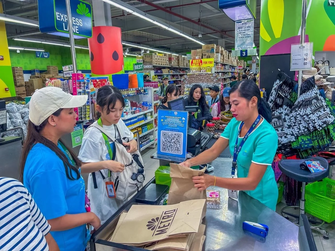 スーパーマーケットのレジカウンターで、店員が商品を袋に詰めている様子が写っています。数人の客が会計を待っており、活気のある買い物の場面です。