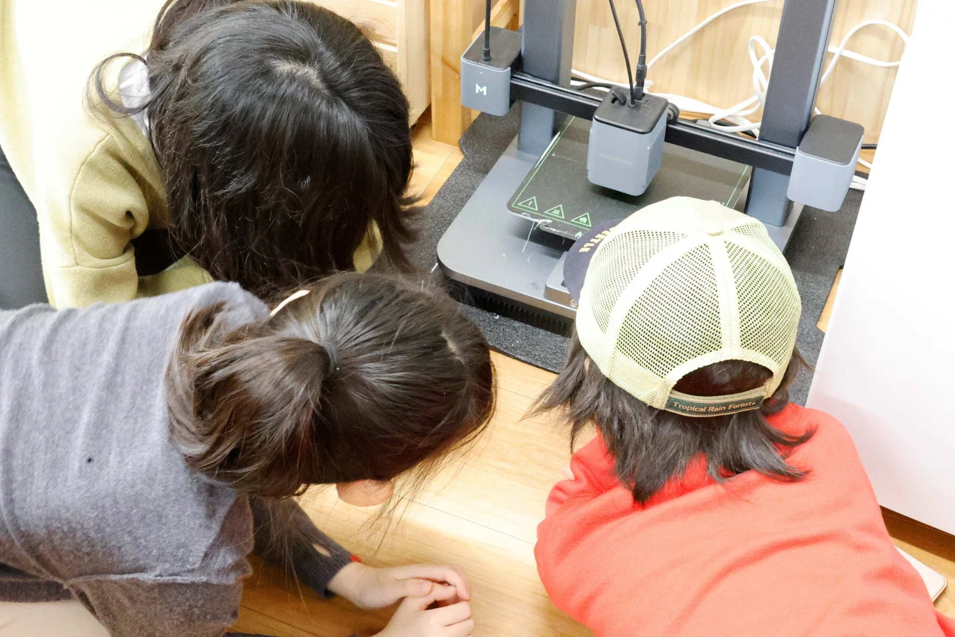 3人の子供たちが3Dプリンターの周りに集まり、その動作を熱心に観察しています。