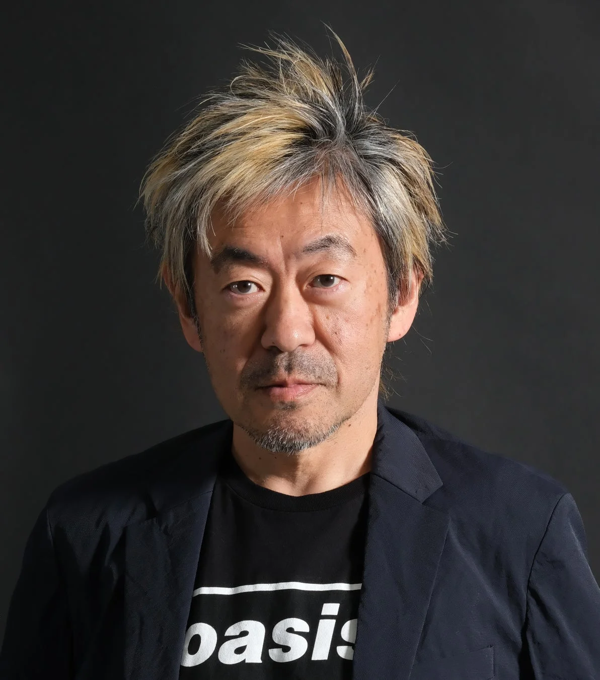 黒い背景の前でカメラをまっすぐ見つめる、ブリーチされた髪とゴアティが特徴的な男性のポートレート