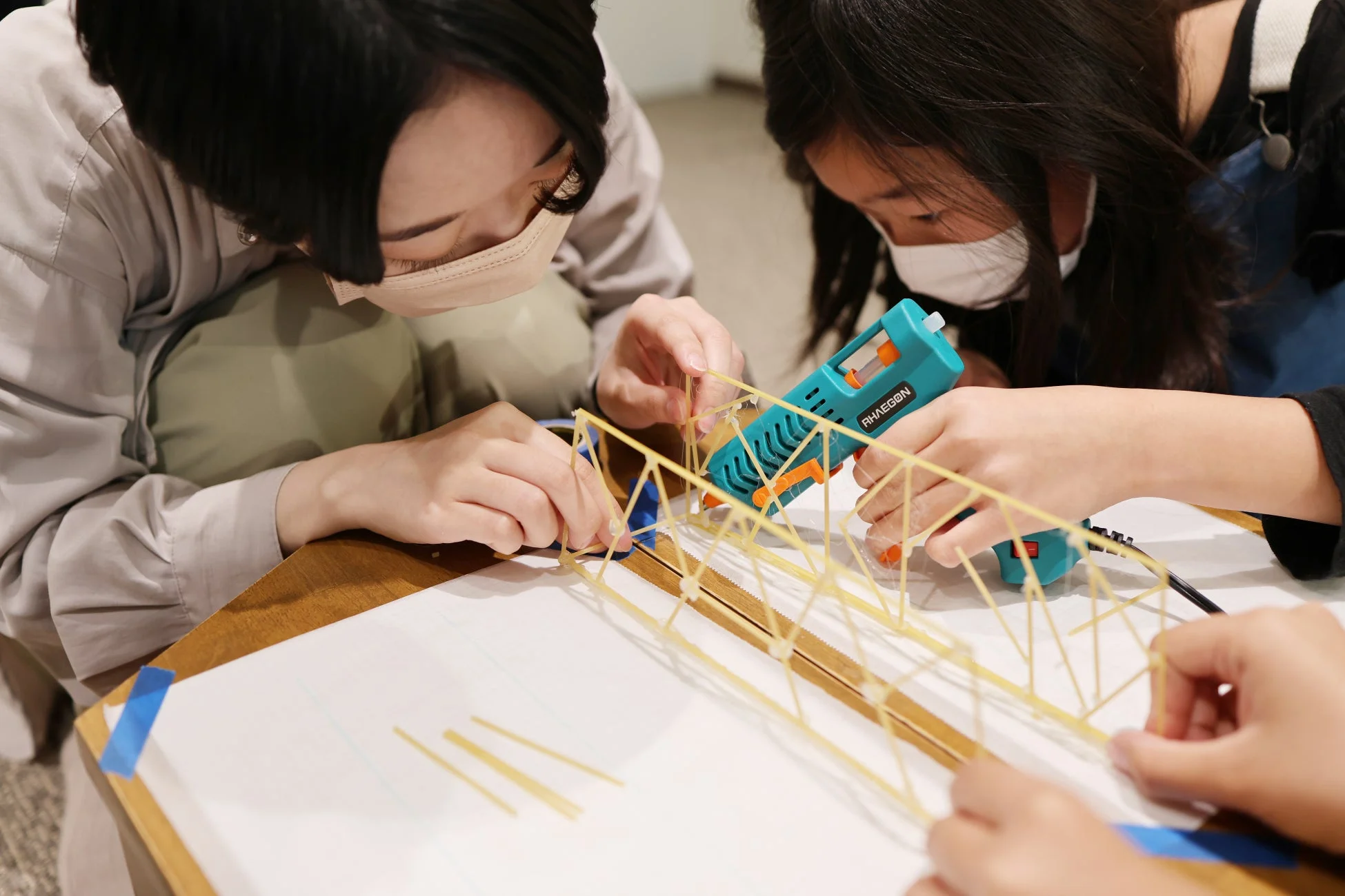 二人の人物がマスクを着用し、スパゲッティとグルーガンを使って橋のような構造物を共同で作成している様子。