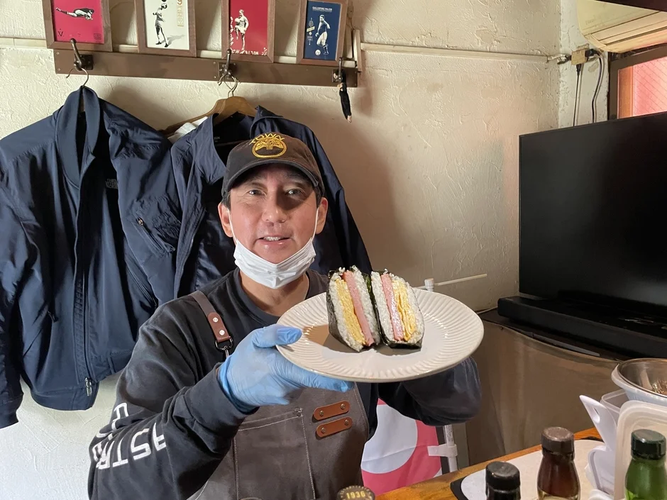 マスクとエプロンを着用した男性が、スパムと卵焼きを挟んだおにぎりを2つ載せた白い皿を差し出している