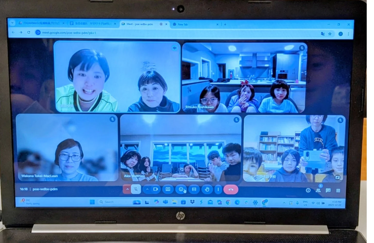 ラップトップPCの画面にGoogle Meetのビデオ会議が表示されています。複数の参加者（大人や子供）が各々の画面に映り、オンラインで交流している様子が伺えます。会議のタイトルや参加者名、日時などの情報も確認できます。